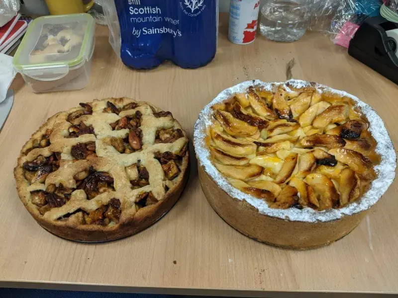 A photo of two cakes, tarts with fruit on top, on a table with containers and drinks behind