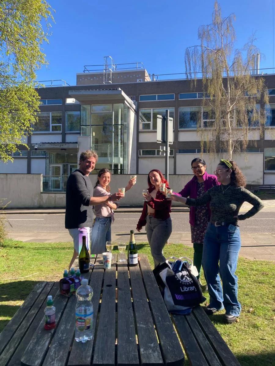 Photo of the group members outdoors standing with raised glasses by a picnic table