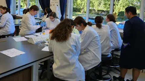 Students in a lab wearing white coats working at benches 