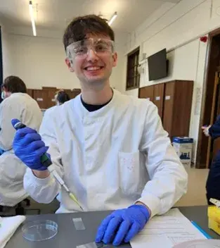 A student, wearing lab coat, safety glasses and gloves, smiles for the camera whilst working at a bench