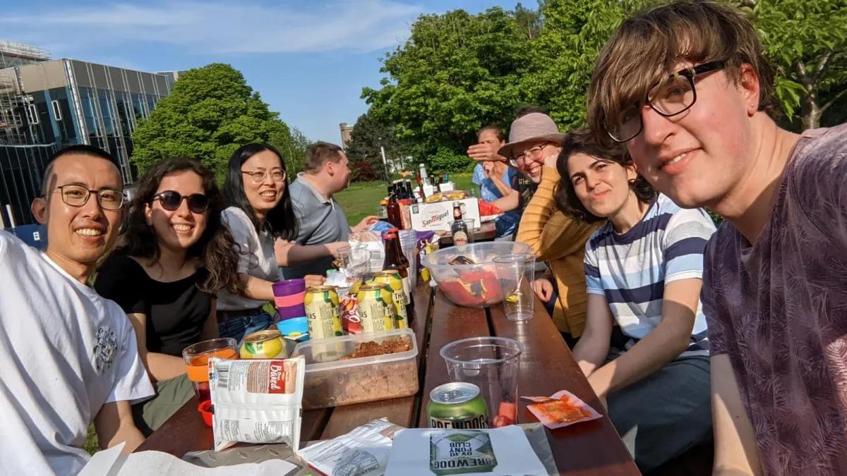 Photo of the lab group members seated outside on a picnic table with food and drinks