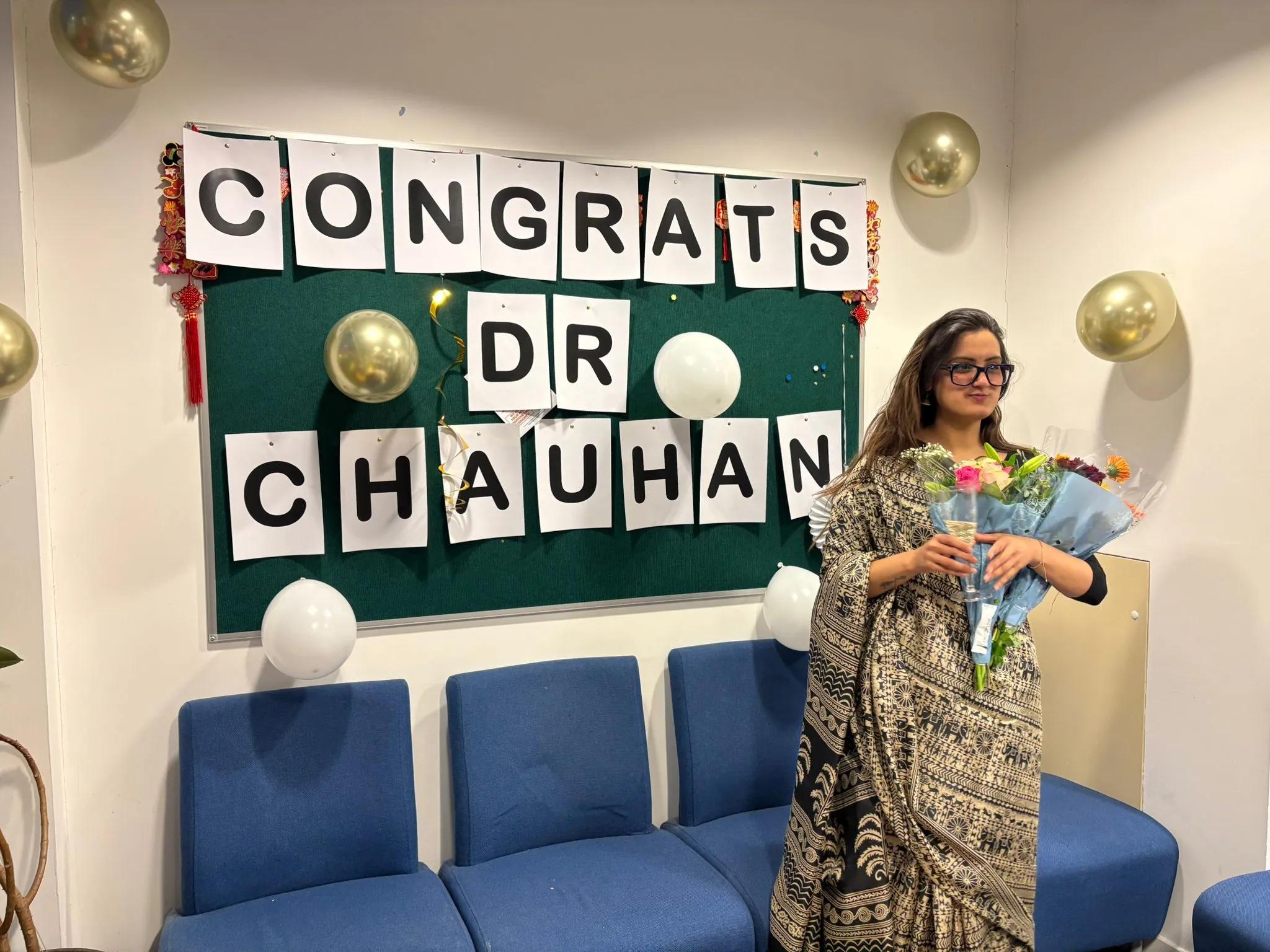 Photo of Mehak holding bunches of flowers in front of a noticeboard with "Congrats Dr Chauhan" printed out on pieces of paper 