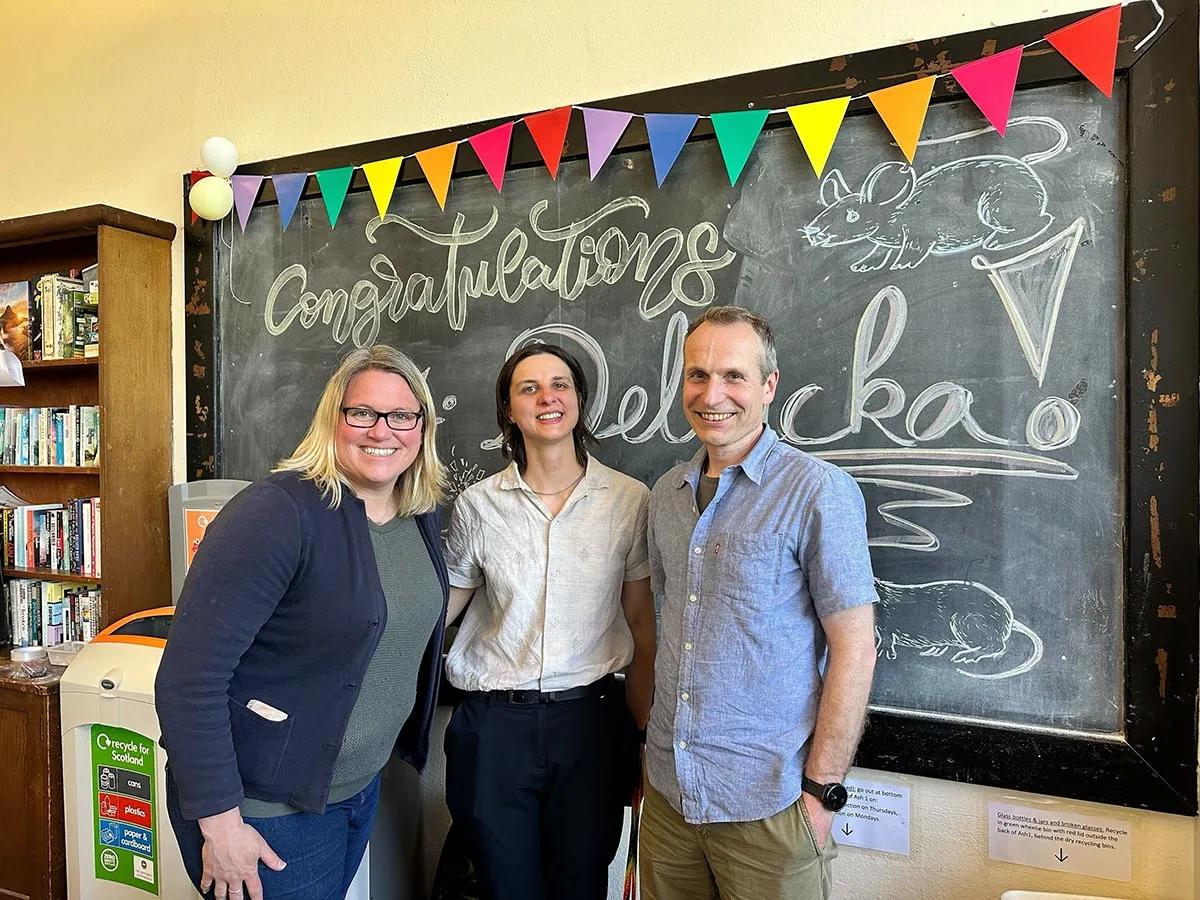 Photo of Agata with two others standing in front of a blackboard with the word 'congratulations' written on it and colourful bunting along the top