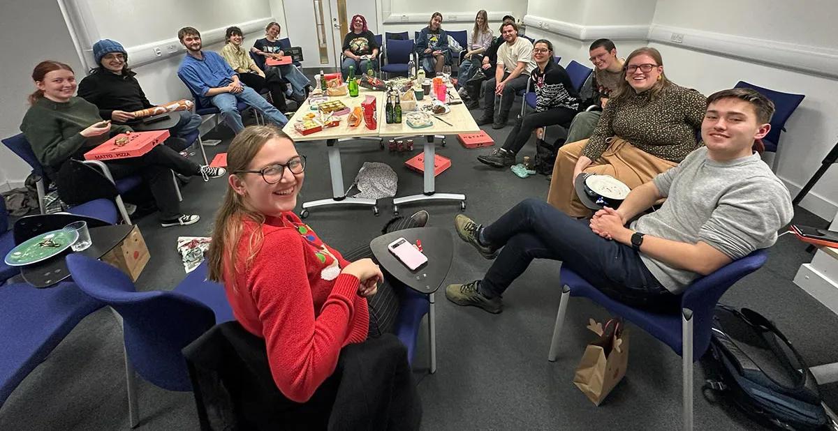 Photo of the lab group members sitting in a circle around tables with pizza boxes and other food and drink around them