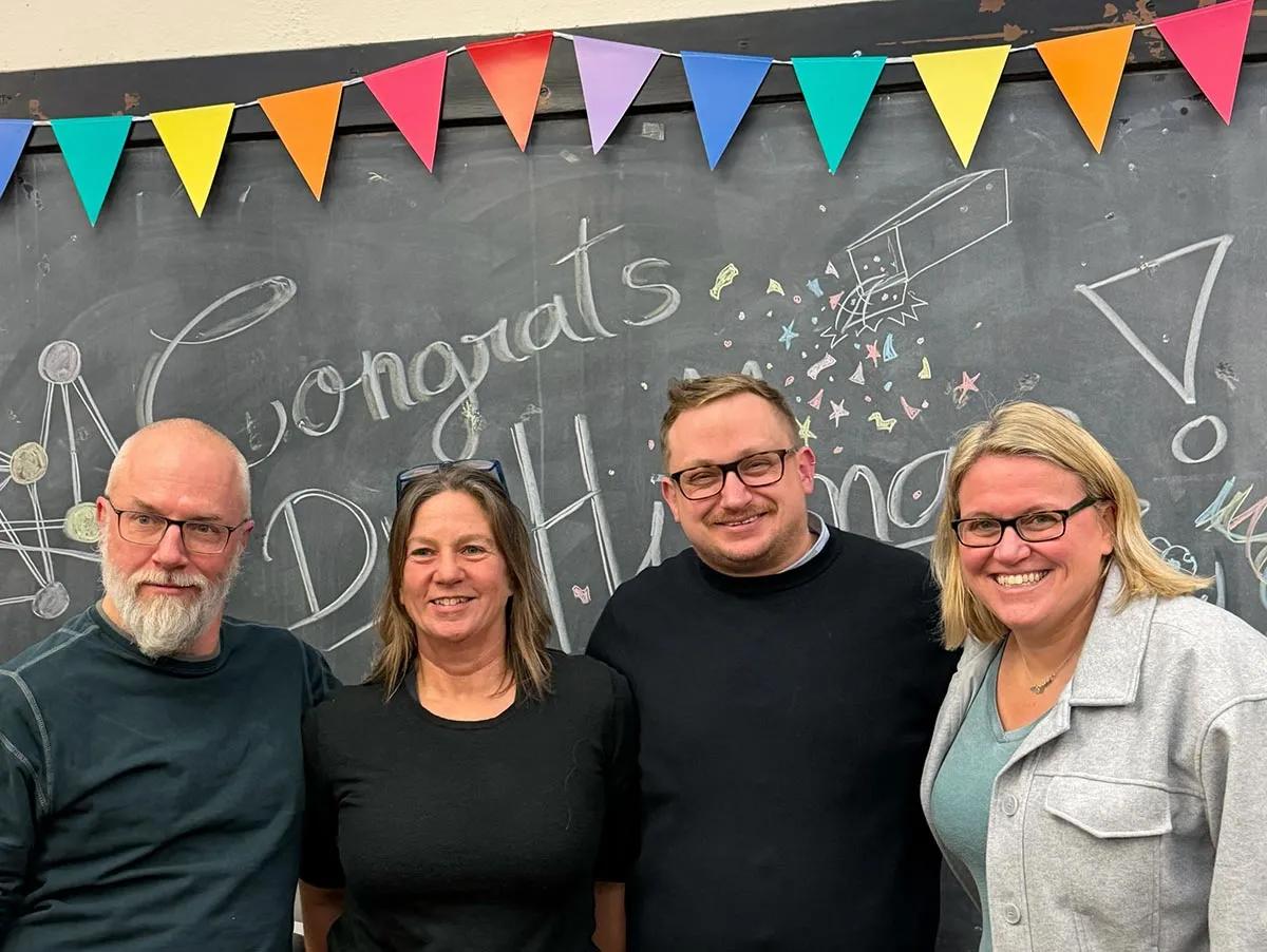 Photo of Sam with three others standing in front of a blackboard with the word 'congratulations' written on it and colourful bunting along the top