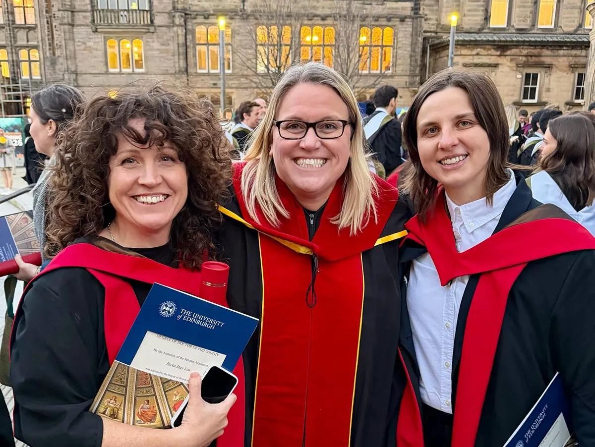 Photo of Rivka and Agata (with Amy) celebrating their graduations in graduation gowns