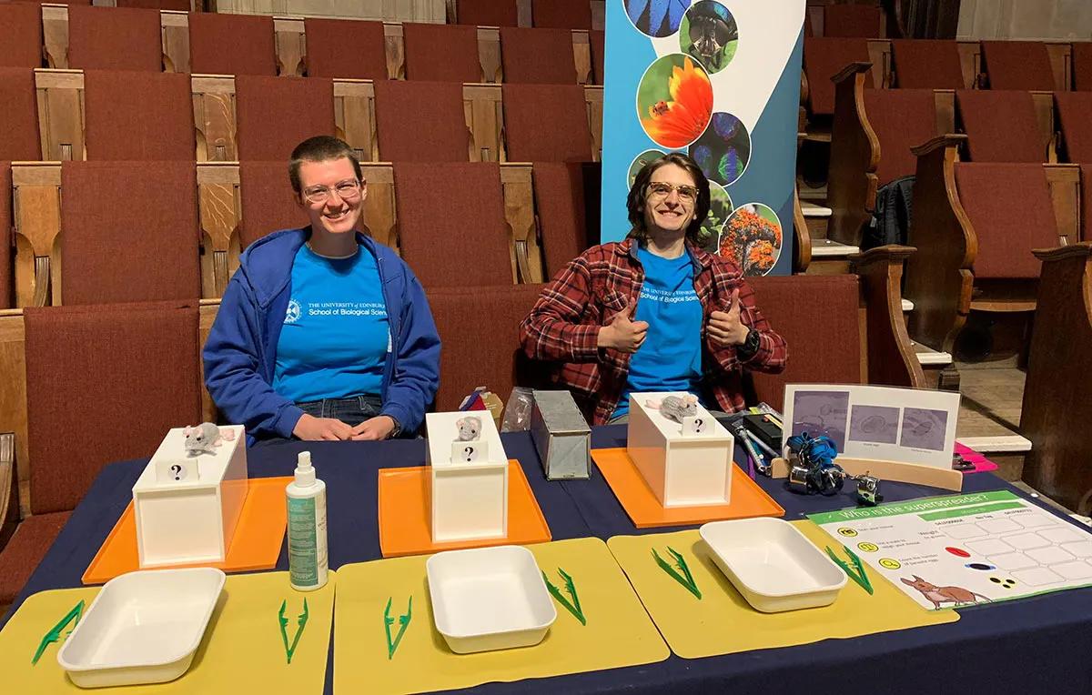 Photo of two lab group member in a lecture theatre witha table set up with props for University of Edinburgh doors open day 2025