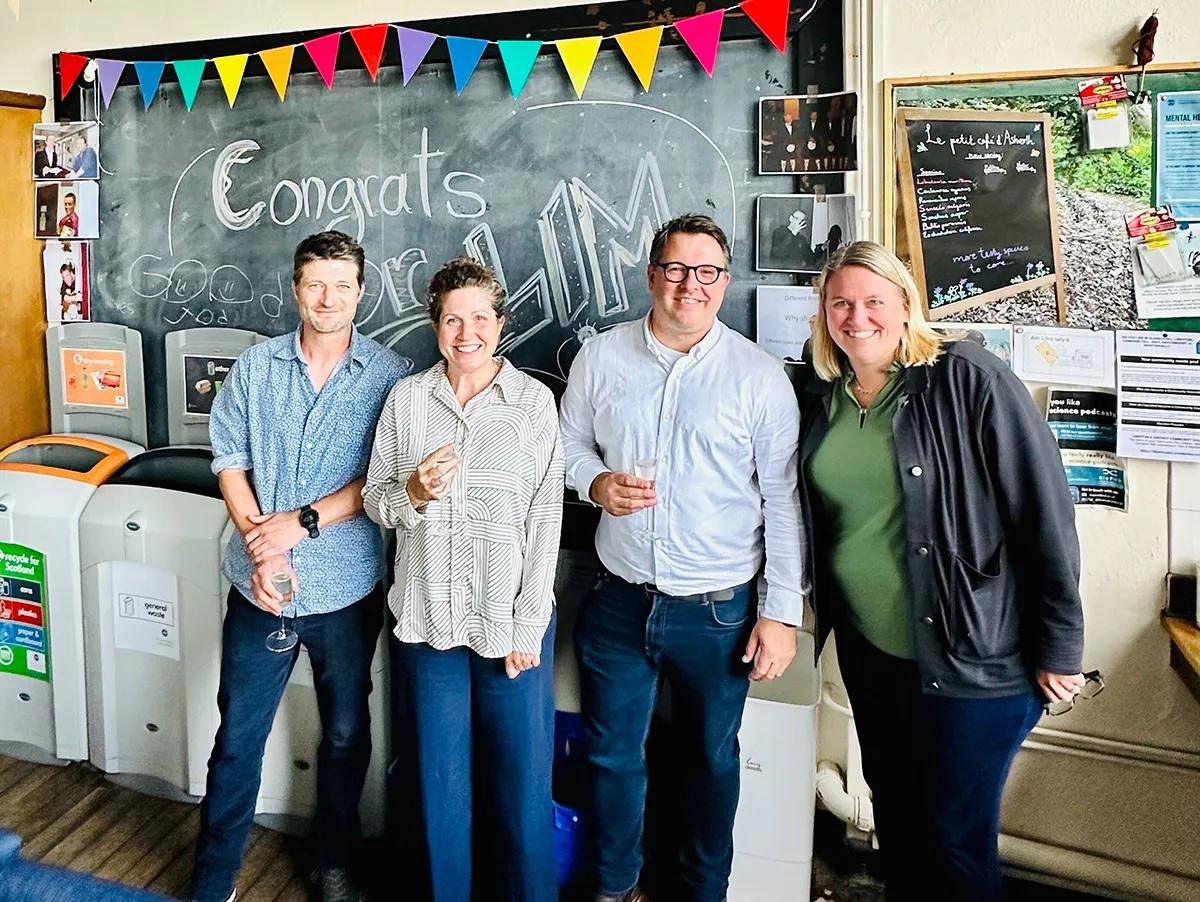 Photo of Rivka with three others standing in front of a blackboard with the word 'congratulations' written on it and colourful bunting along the top