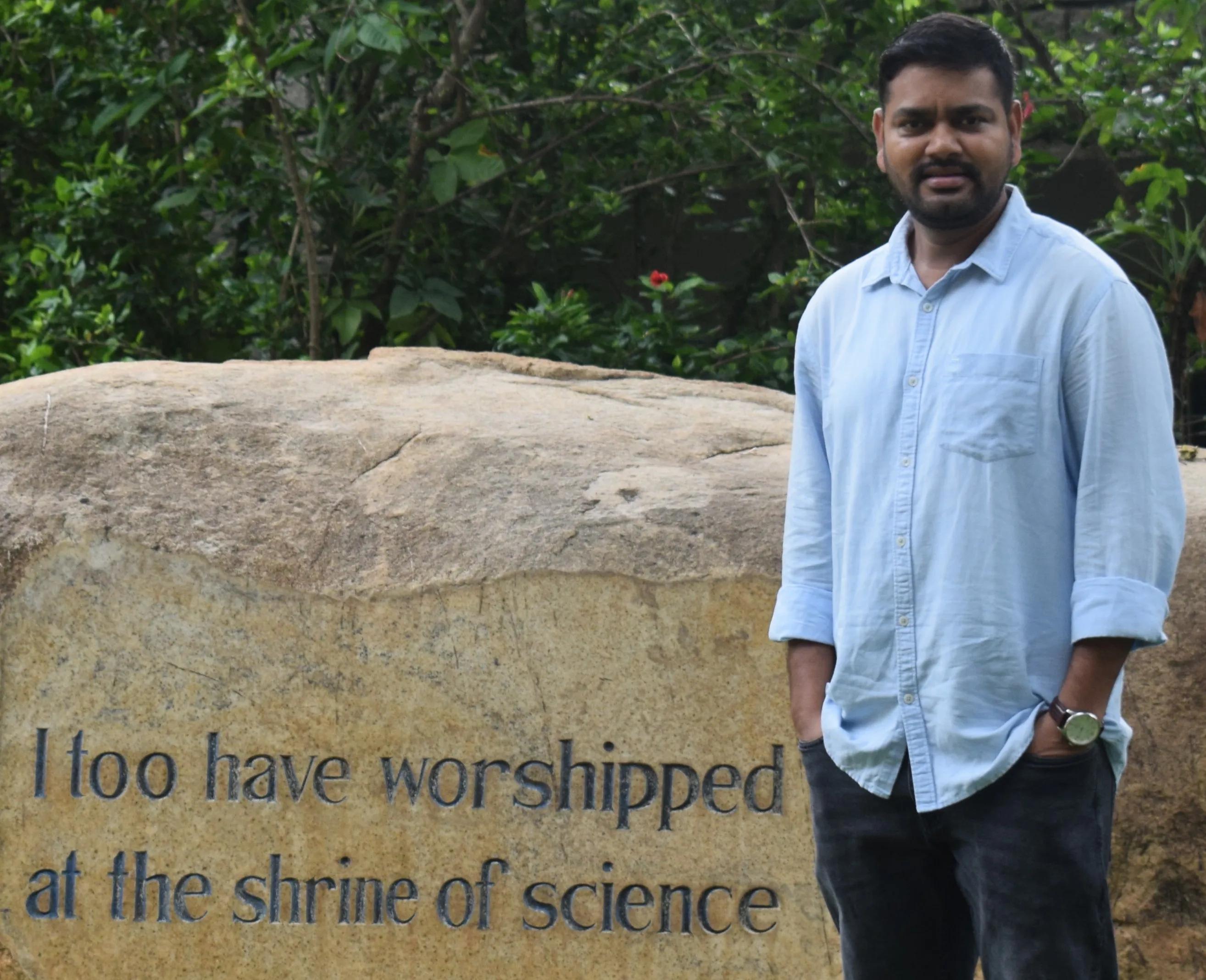 Image of Hashim Reza standing in front of a boulder with the words "I too have worshipped in the shrine of science" inscribed