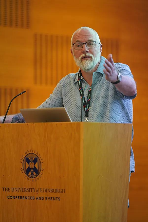 Photo of Professor Tom Little speaking at a wooden podium branded with the University's logo