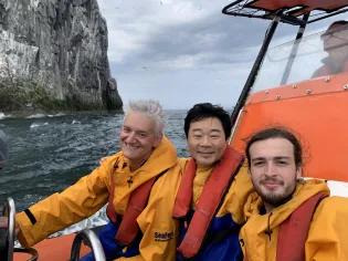 Photo of Justin, Moto Ashikari and Nathan Lacombe on a boat in yellow coats and life jackets, in front of Bass Rock