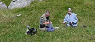 Photo of 2 researchers doing fieldwork, sitting in a grassy area with grey rocks close by