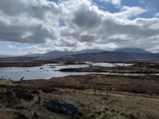 Photo of a landscape with a grassy moore and a body of water in the foreground and hills in the background, the sky above is mostly cloudy