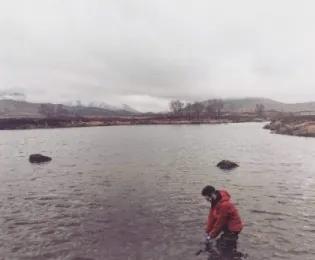 Photo of a lab memeber in a red jacket sitting by a water's edge surrounded by scrubby grass, ready to collect a sample