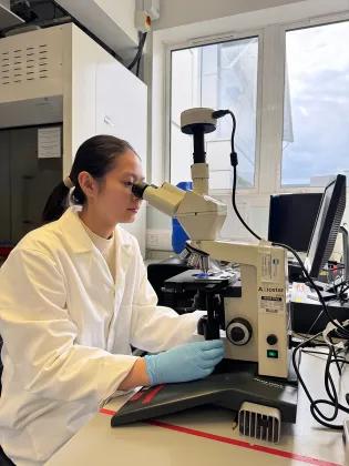 Photo of a student in the lab, wearing lab coat and gloves, using a microscope