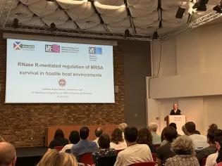 Photo of Gabriele standing behind a podium speaking at the Regulating with RNA in Bacteria & Archaea conference