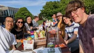 Photo of the lab group members seated outside on a picnic table with food and drinks