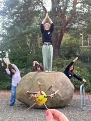 Students pose for photo outside, one stands on a rock with hands up, others behind the rock, and in the foreground, a hand holds an apple with twigs sticking out of it