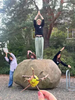 Students pose for photo outside, one stands on a rock with hands up, others behind the rock, and in the foreground, a hand holds an apple with twigs sticking out of it
