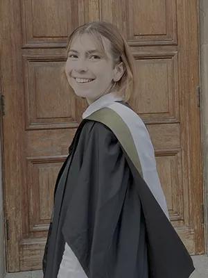 Photo of Olivia in graduation gown, standing in front of a wooden door