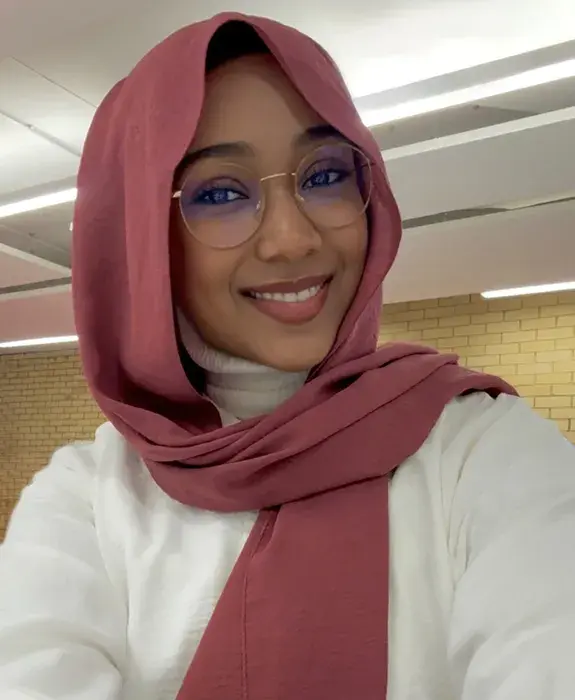 A woman with a white blouse and pink head covering, she has round glasses and is smiling at the camera.