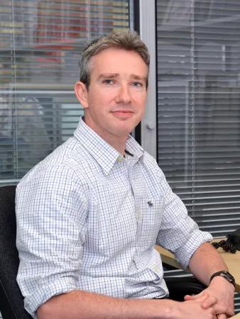 Photograph of a person sitting in an office chair at a desk with blinds and office equipment in the background.