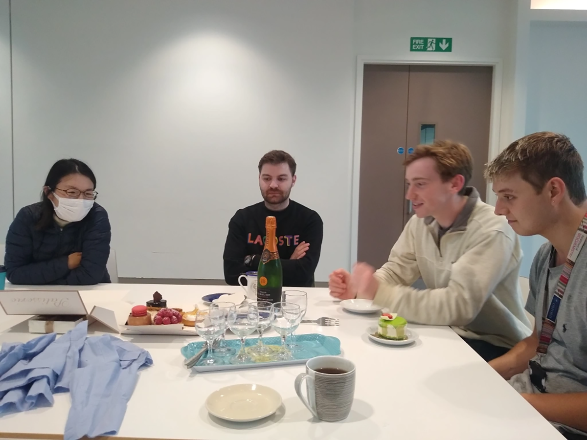 Photo of lab group members seated around a table that has pastries, glasses and a bottle on it