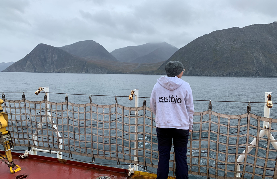 A student in an EASTBIO hoodie stands on the side of a boat looking at a mountainous shoreline