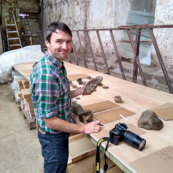 Sandy Hetherington standing in front of a table with fossils, holding a fossil in left hand