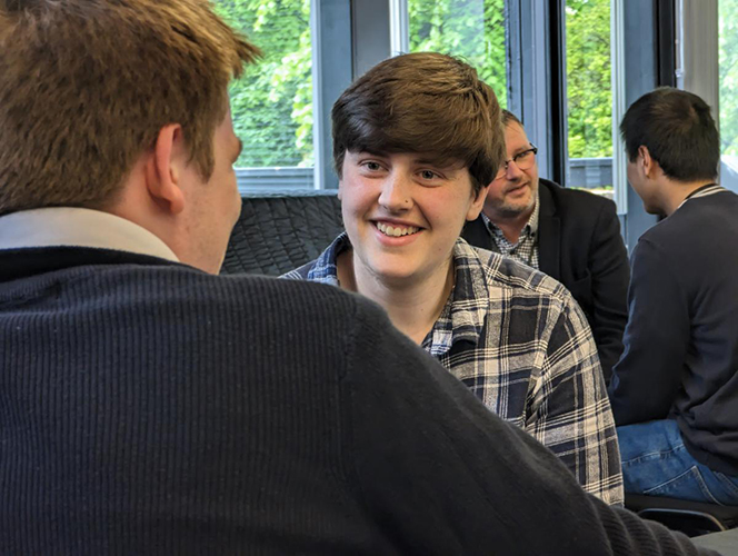 Students and supervisors sit and talk, a smiling student is in foreground