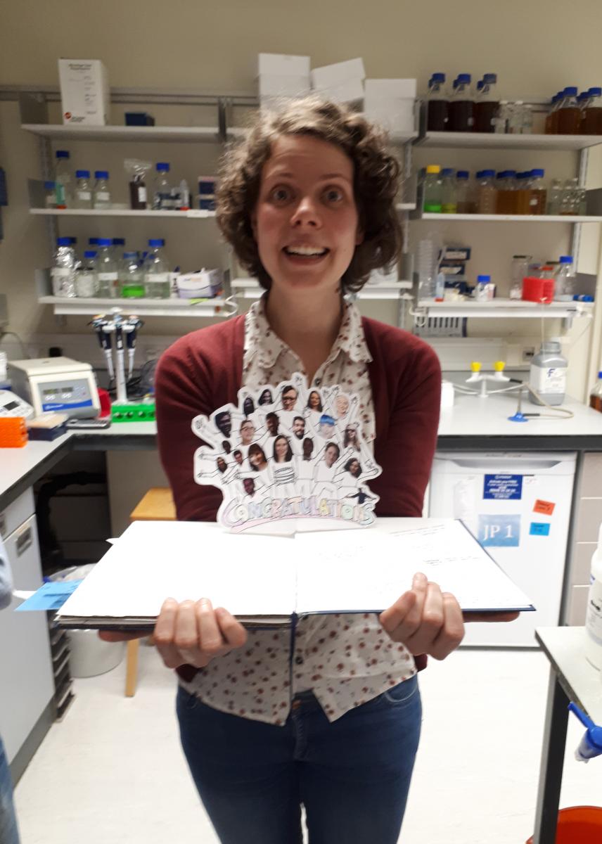 Jovana poses in the lab with an open book and a card with photos of the lab group members on it