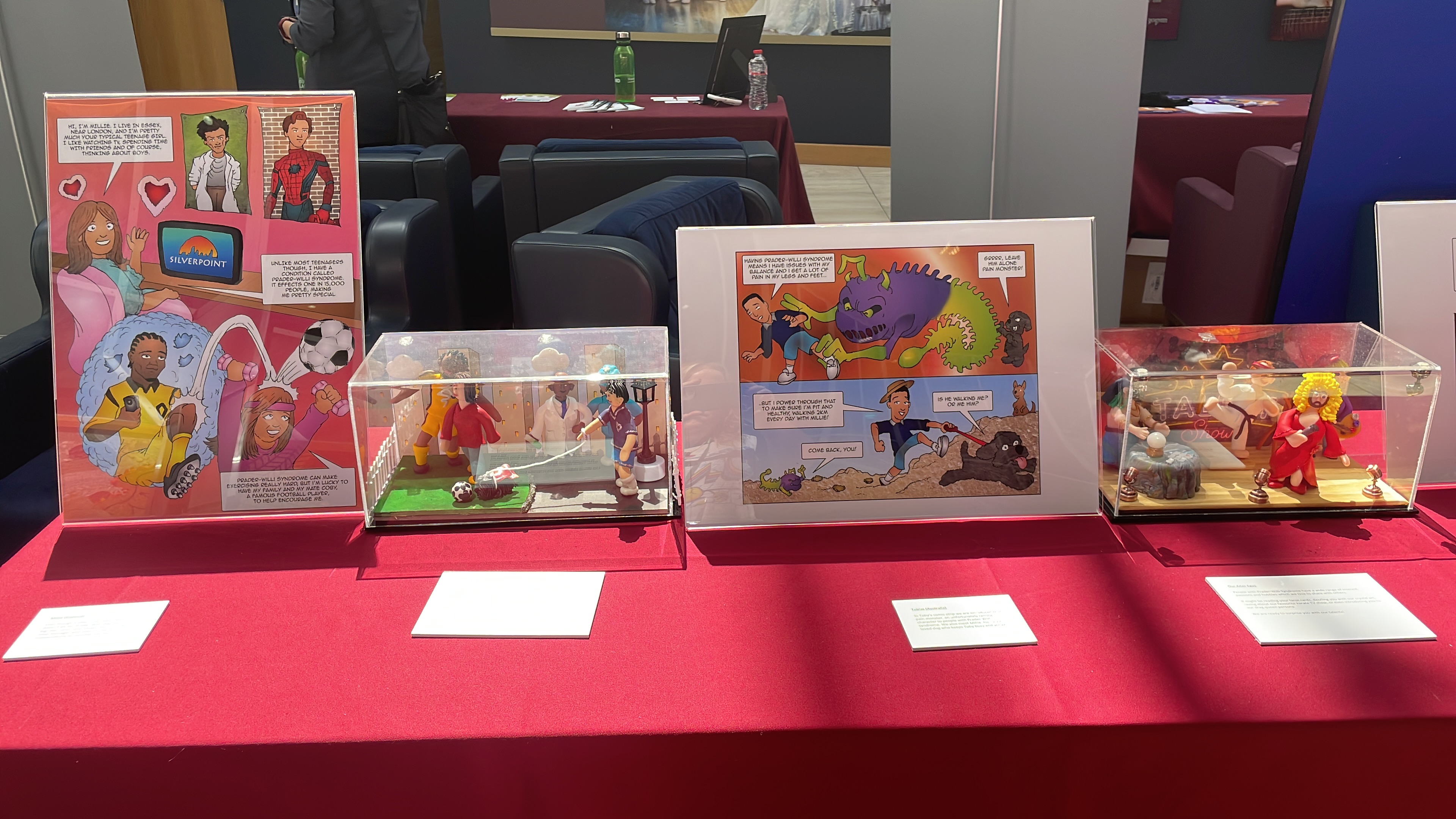 Photo of table at the exhibition, covered in a red cloth and displaying illustrations and figures in glass cases from the comics