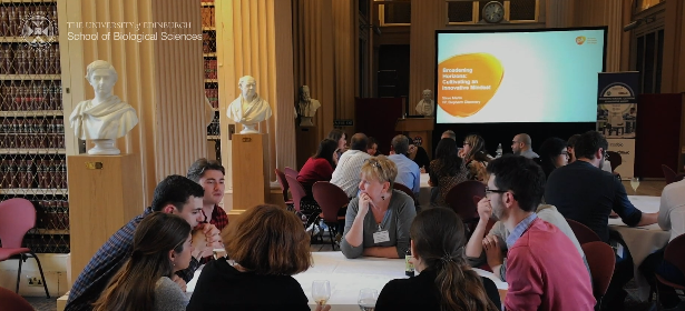 Photo capture from video, showing people sitting around tables talking, and a video screen in the background