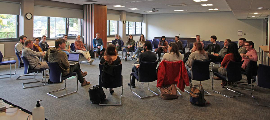 Meeting of students and academics, they sit on chairs arranged in a circle in a large room with windows along left side